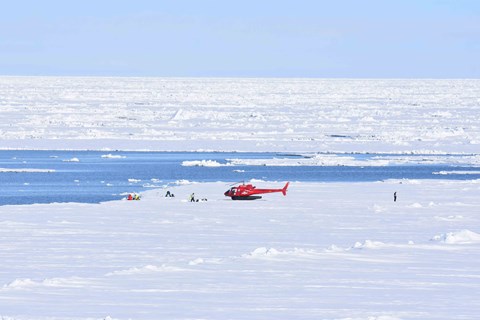 A red helicopter stands on the snow. Open water can be seen in the background.