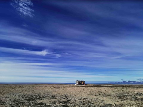 A gray little wooden cabin in a mighty open environment. The ground is covered in gravel and the sky is blue.