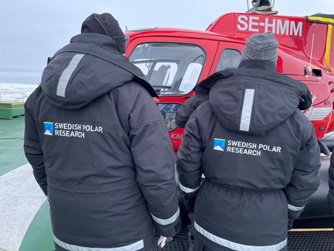 Två personer står med ryggen vänd mot kameran. På jackan står det Swedish Polar Research. 
