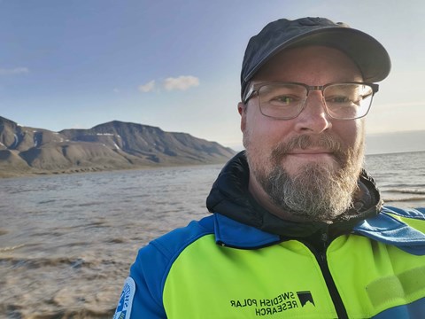 Portrait of Andreas Johnsson. He has a yellow and blue jacket, a cap, glasses and a beard. In the background you can see water and mountains.
