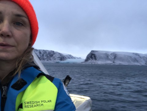 Kvinna med röd mössa och gul och blå jacka. I bakgrunden syns vatten, berg och glaciärer.