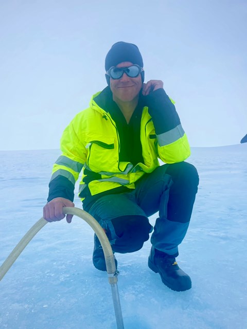 En man i gul jacka och solglasögon sitter hukad på isen.