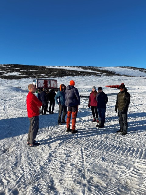 Ett antal personer står ute på snön och pratar med varandra medan solen skiner. 