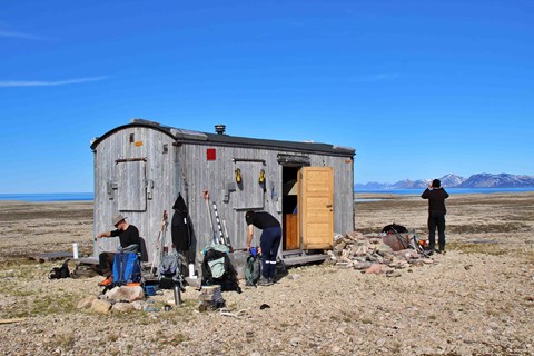 The researchers next to a gray wooden cabin.