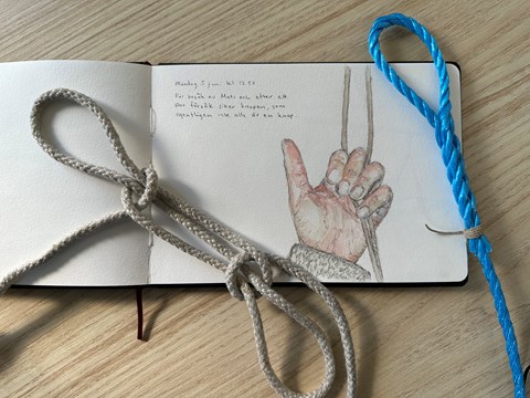 Teckning/akvarell av en hand som håller ett rep. Vid sidan av teckningen ligger ett beige och blått rep.