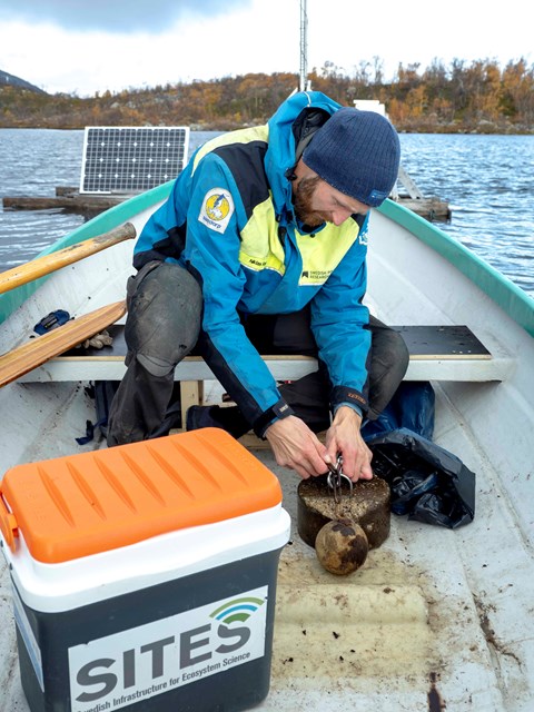 A man in a blue and yellow jacket sits in a boat bent over a metal weight