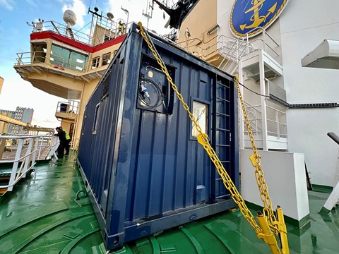 En blå container står stadigt förankrad med gula kättingar i det gröna fartygsdäcket. 