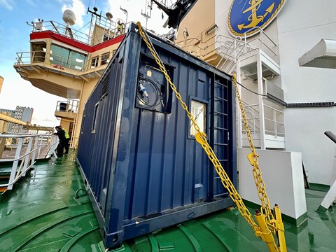 En blå container står stadigt förankrad med gula kättingar i det gröna fartygsdäcket.