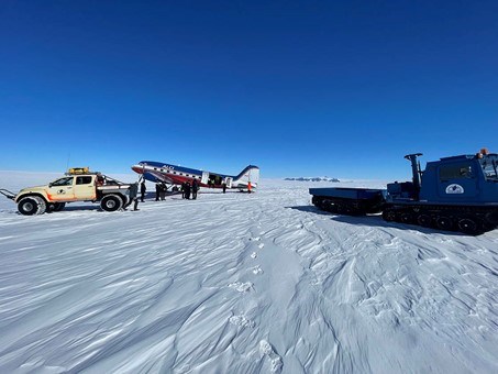 Ett fraktplan har landat på isen. Framför planet står en bil och en bandvagn. 